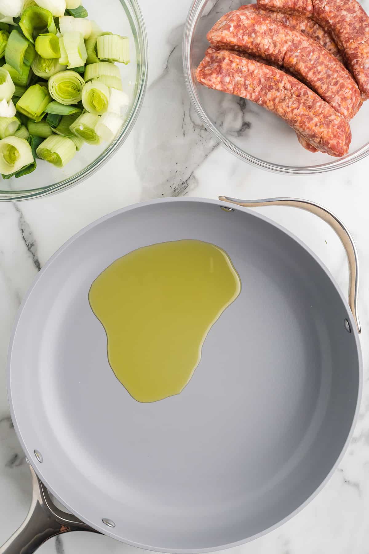 A skillet with olive oil sits on a marble counter near bowls of chopped leeks and raw sausage links.