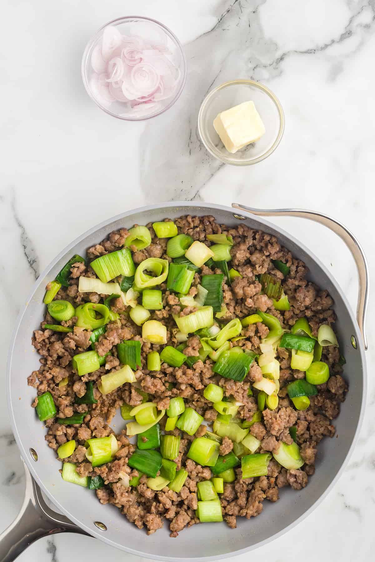 Top view of a skillet with cooked ground meat and chopped green onions, beside bowls of butter and sliced shallots.