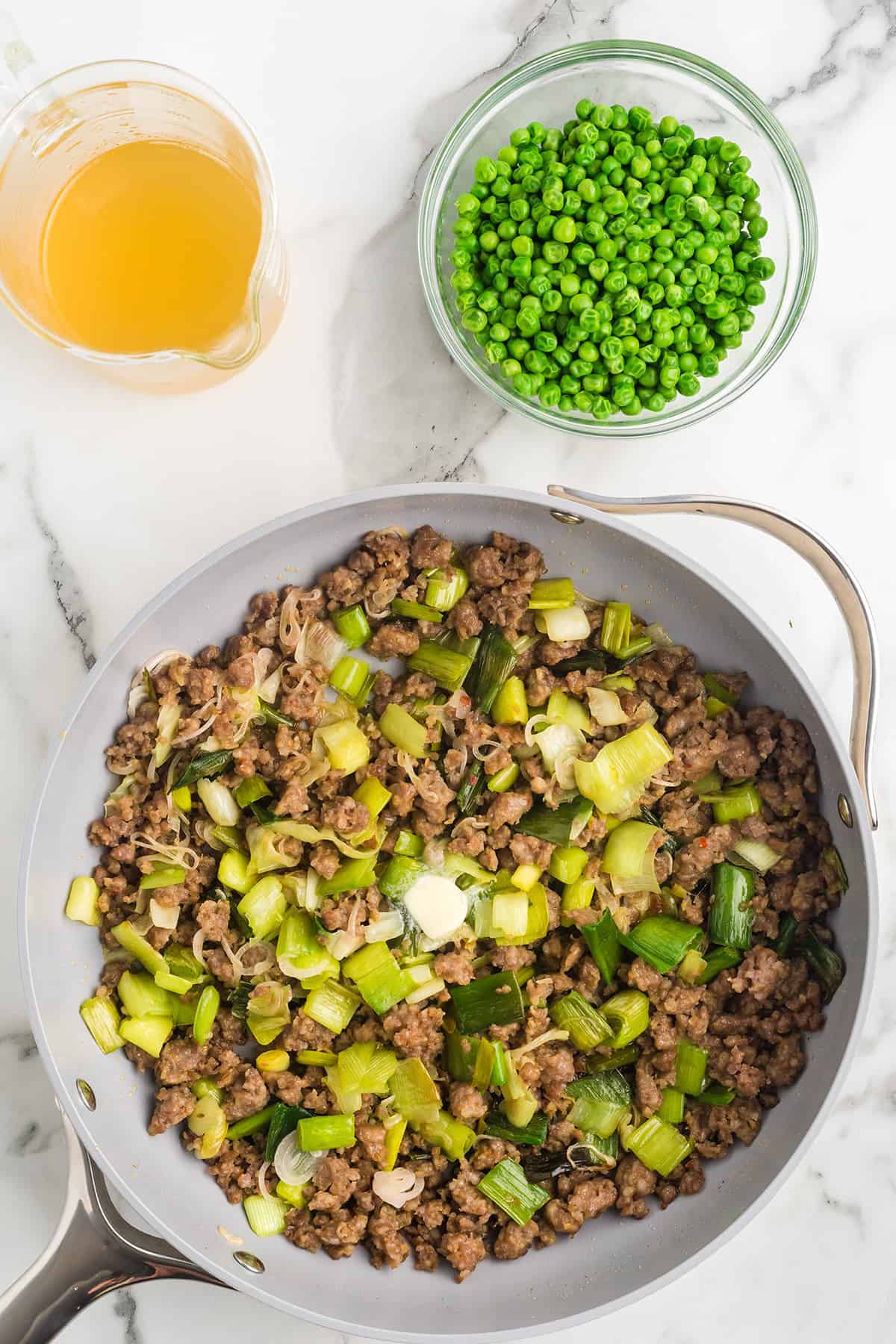 A skillet with cooked ground meat and green vegetables, next to a bowl of peas and a cup of broth on a marble counter.