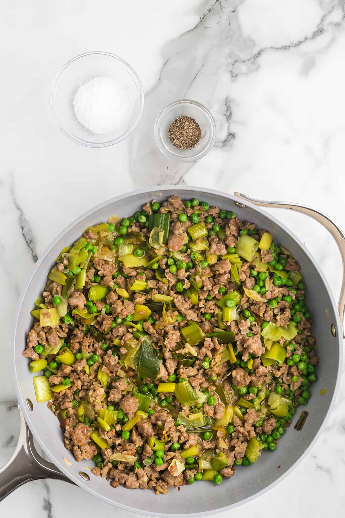 A skillet with ground meat, peas, and leeks on a marble counter, next to bowls of salt and pepper.