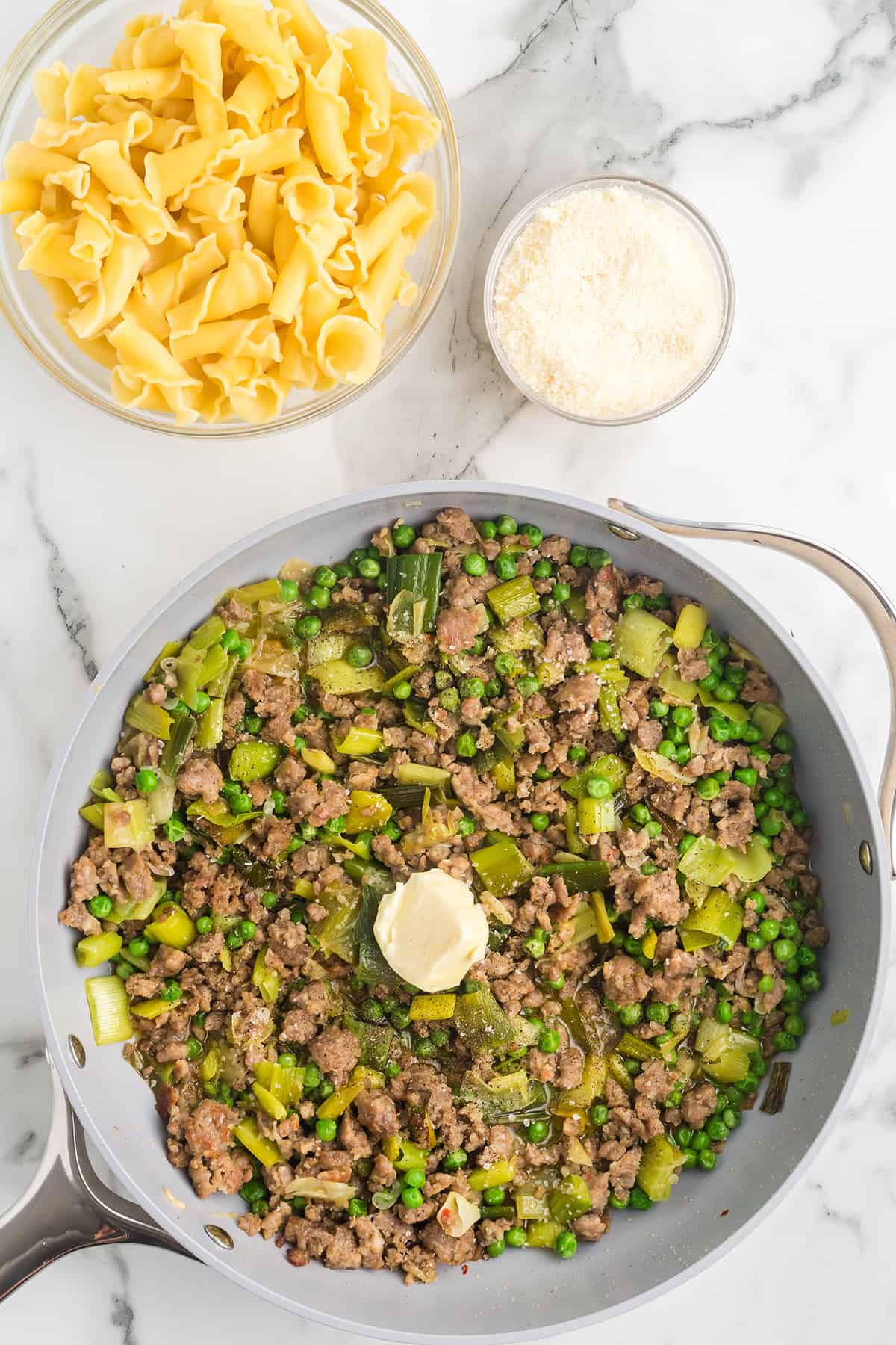 Cooked pasta, grated cheese, and a skillet with ground meat, peas, leeks, and a dollop of butter on a marble surface.