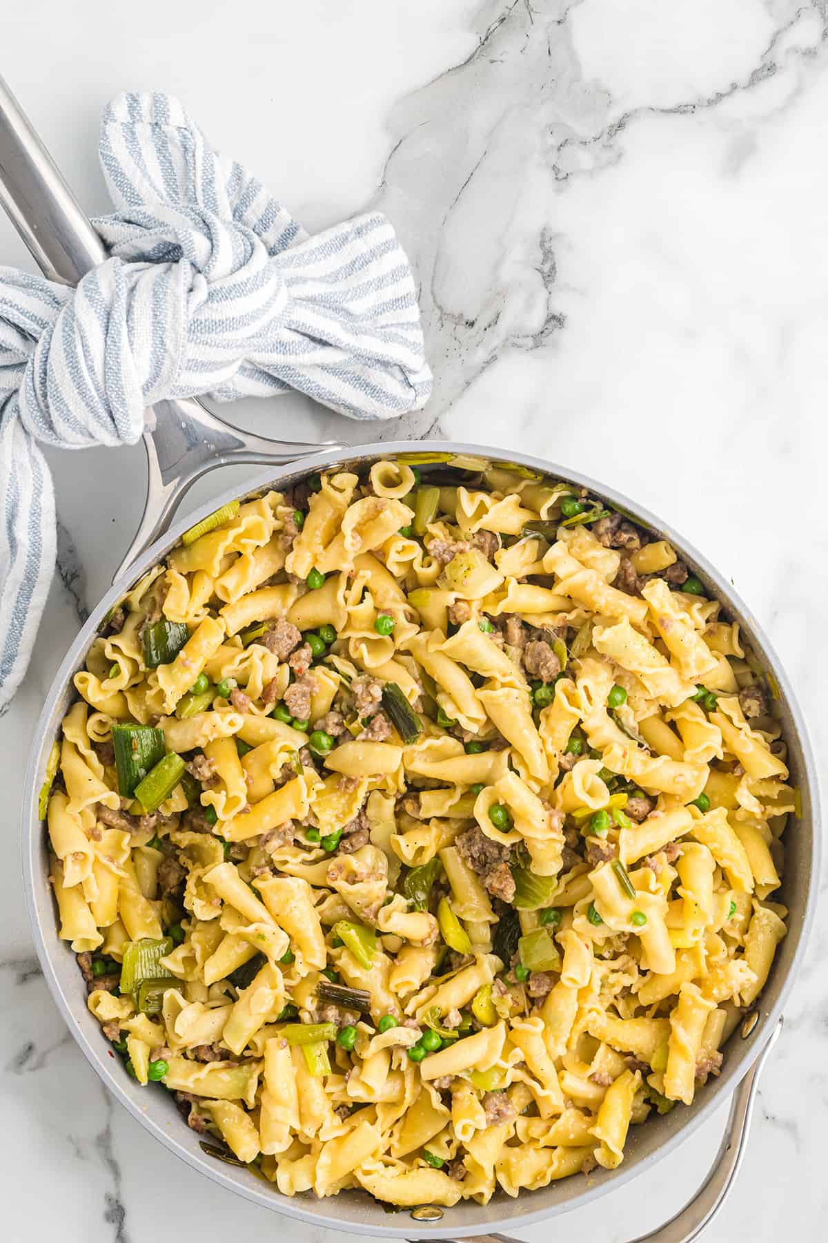 A skillet filled with pasta, ground meat, green vegetables, and a striped towel wrapped around the handle.