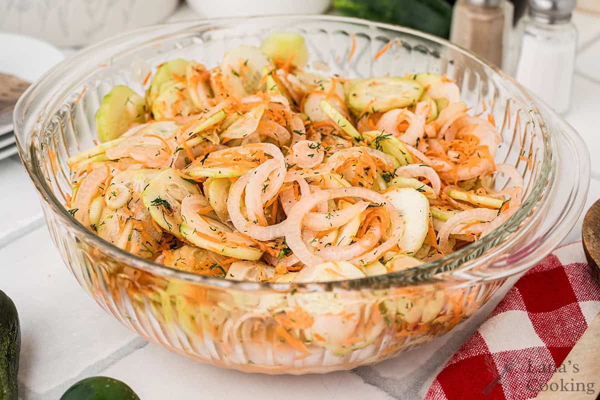 A glass bowl filled with a salad made of sliced cucumbers, onions, and shredded carrots, seasoned with herbs and spices.