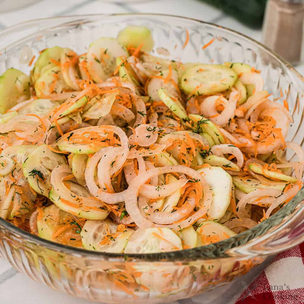 A glass bowl filled with sliced cucumbers, onions, and shredded carrots mixed with herbs, sitting on a table.