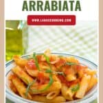 A bowl of penne pasta arrabbiata topped with tomato sauce and fresh basil, next to a bottle of olive oil.