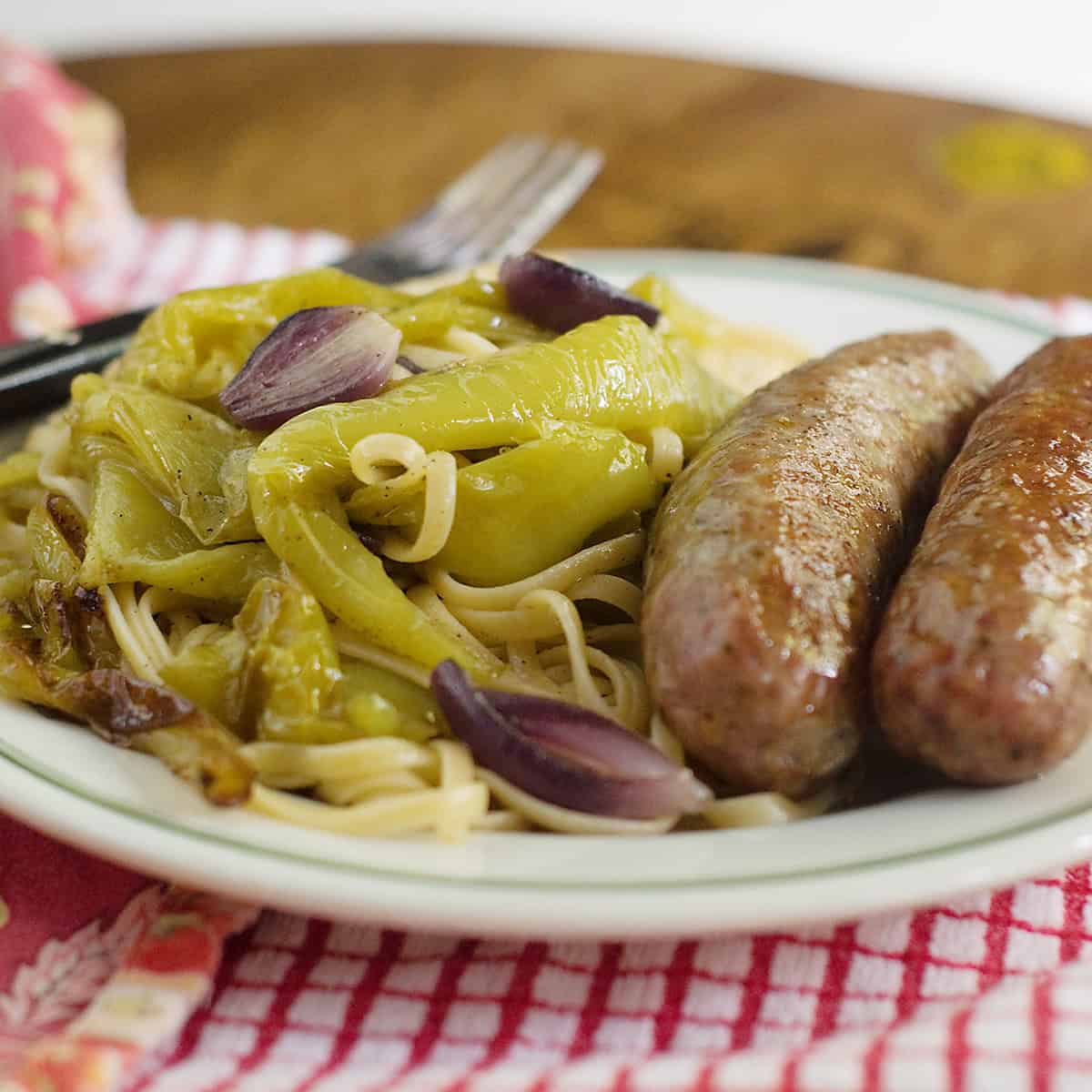 Plate of pasta with roasted green peppers, red onions, and two sausages on a table with a fork and napkin.