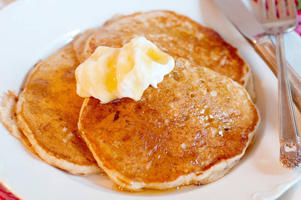 Two pancakes topped with a dollop of butter and drizzled with syrup on a white plate, with a fork beside them.