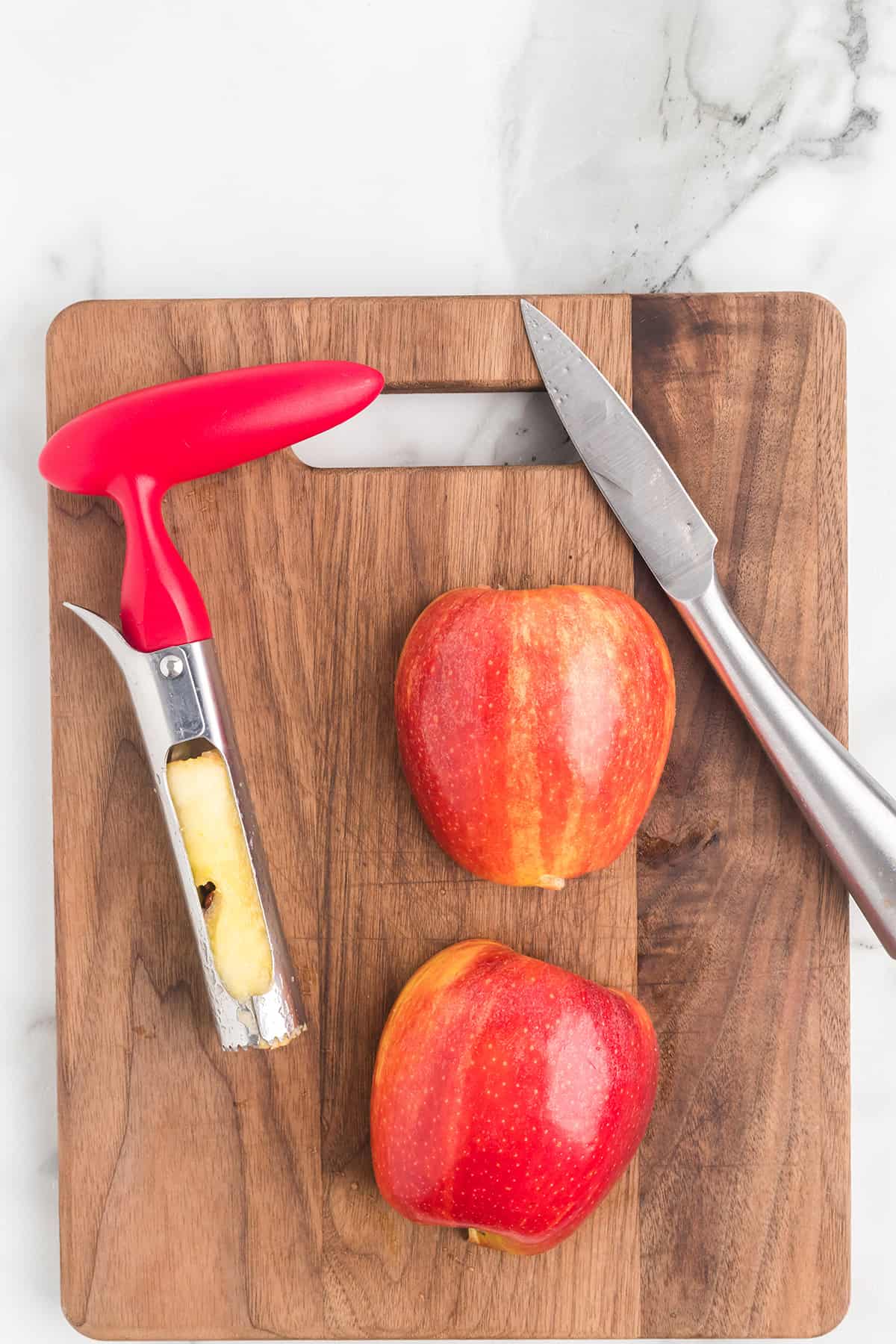 A cored and halved apple, corer, and knife on a wooden cutting board with a marble background.