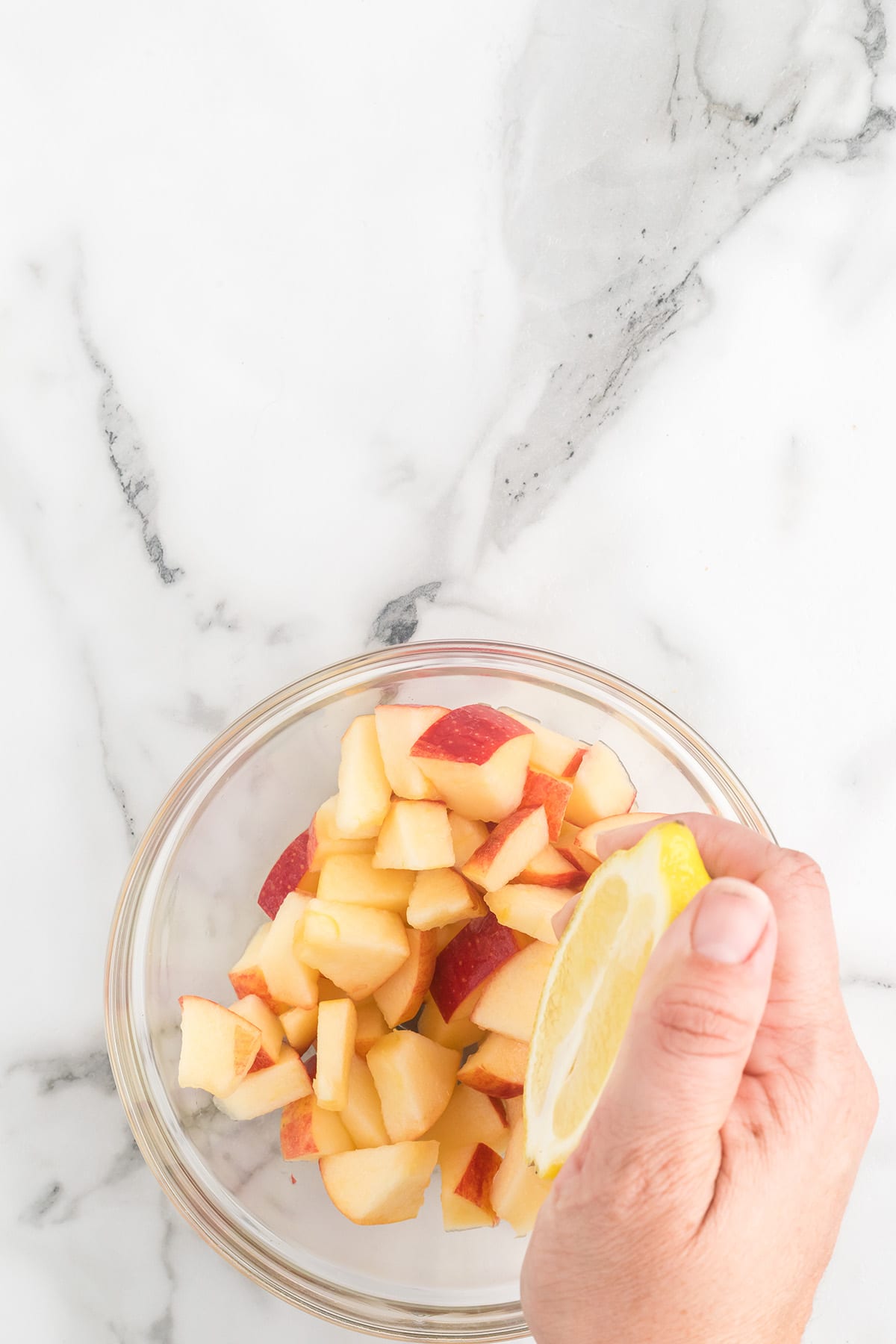 Hand squeezing lemon juice over a bowl of chopped apples on a white marble surface.
