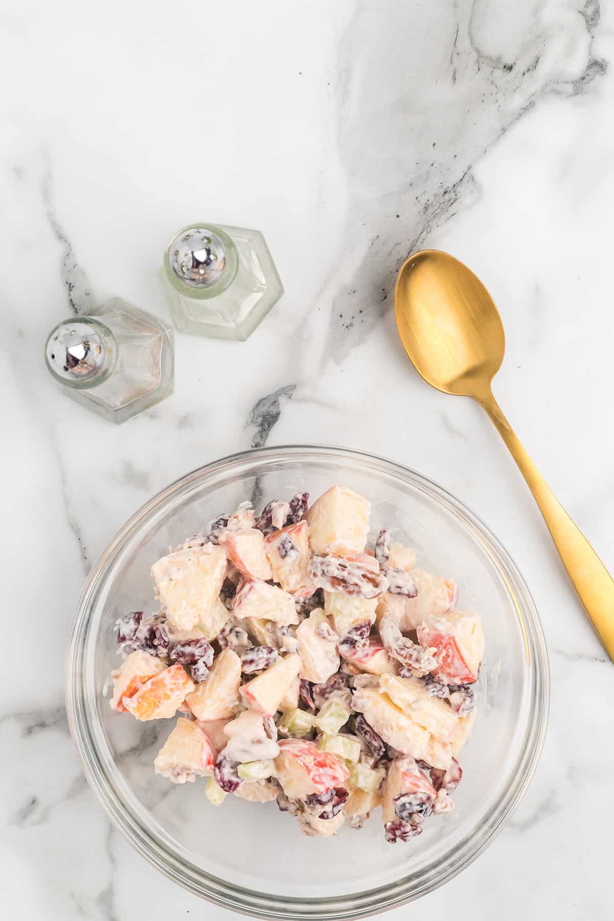 A glass bowl of fruit and nut salad with a gold spoon and salt and pepper shakers on a marble surface.