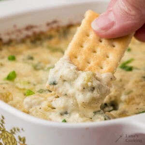 A hand dips a rectangular cracker into a creamy, cheesy baked dip in a white dish.