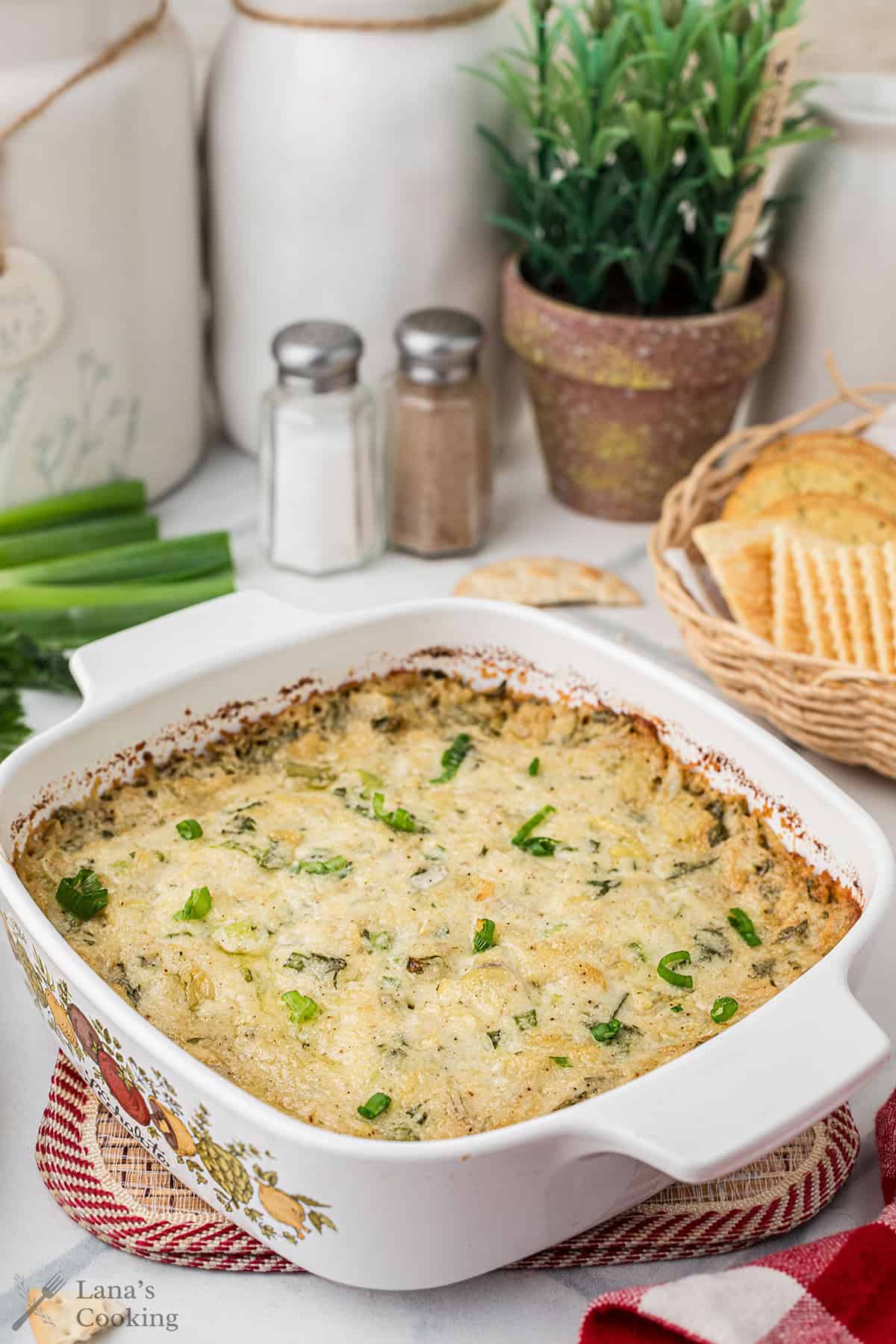 Hot artichoke dip topped with green onions in a white dish, with crackers and seasoning in the background.