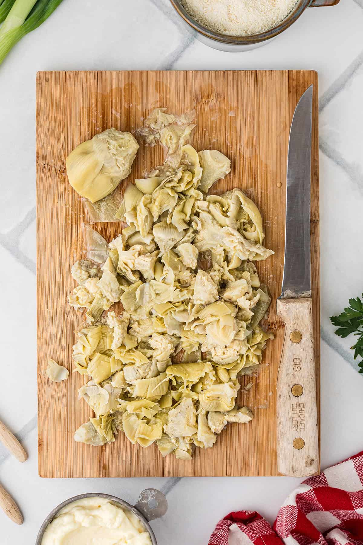 Chopped artichoke hearts on a wooden cutting board with a knife and various ingredients nearby.