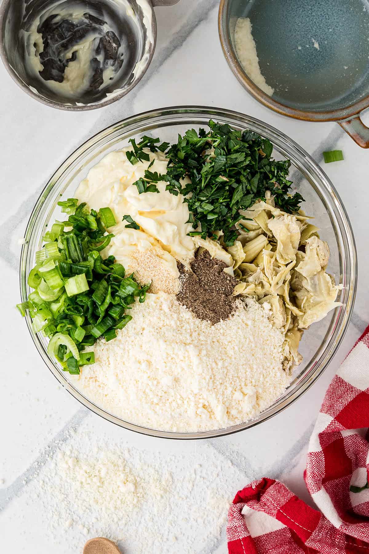 Glass bowl with chopped green onions, parsley, artichokes, grated cheese, mayo, and pepper, on a white surface.