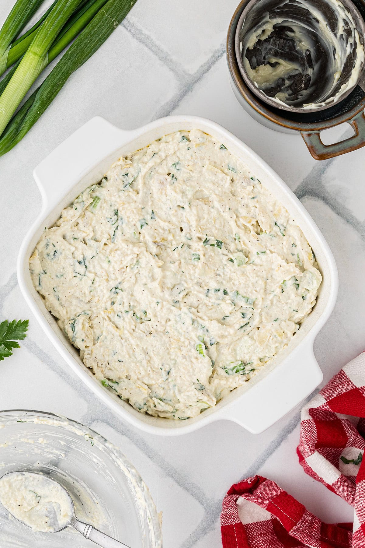 Square white dish filled with creamy dip mixture, surrounded by green onions and a red kitchen towel.