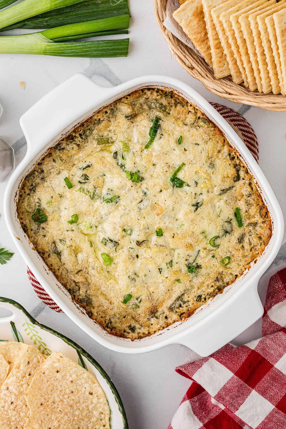 A baked spinach and artichoke dip in a white dish, surrounded by crackers and green onions.