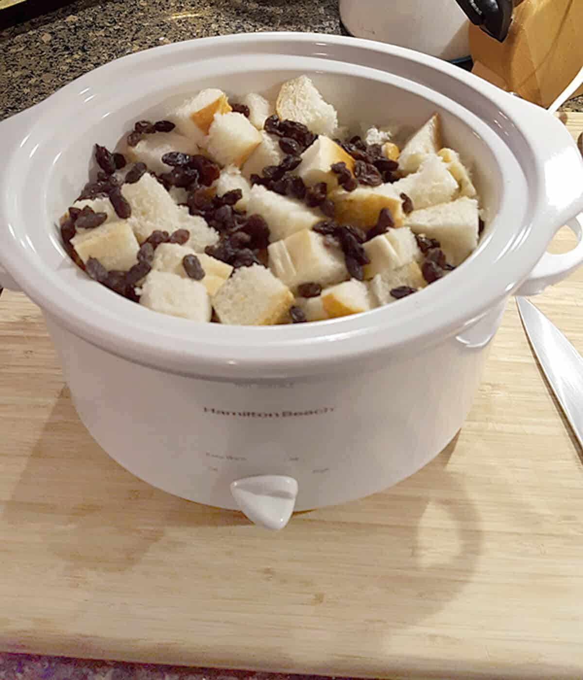 Slow cooker filled with cubed bread and raisins sits on a wooden cutting board next to a knife.