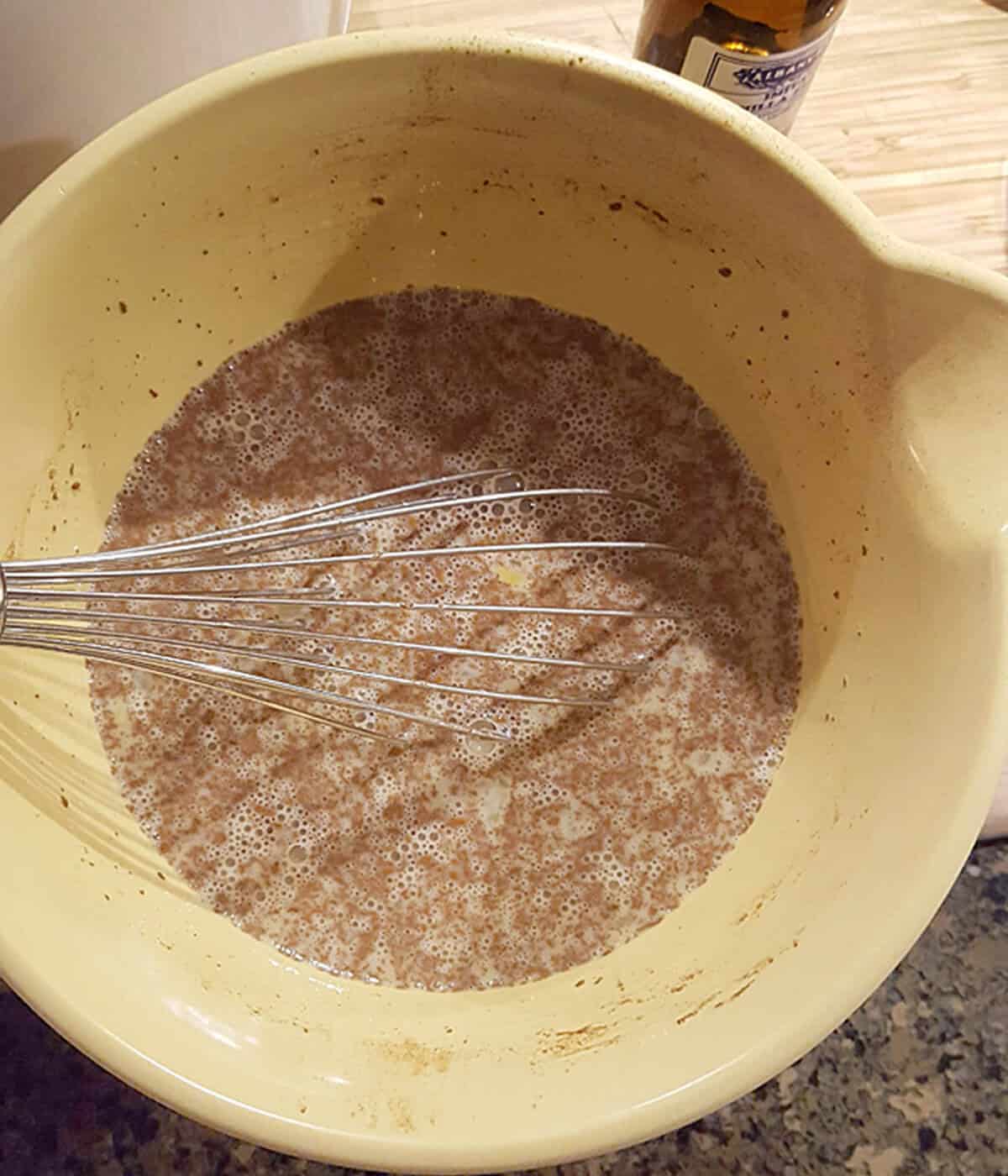 A metal whisk in a yellow bowl mixing a brown, frothy liquid on a kitchen counter.