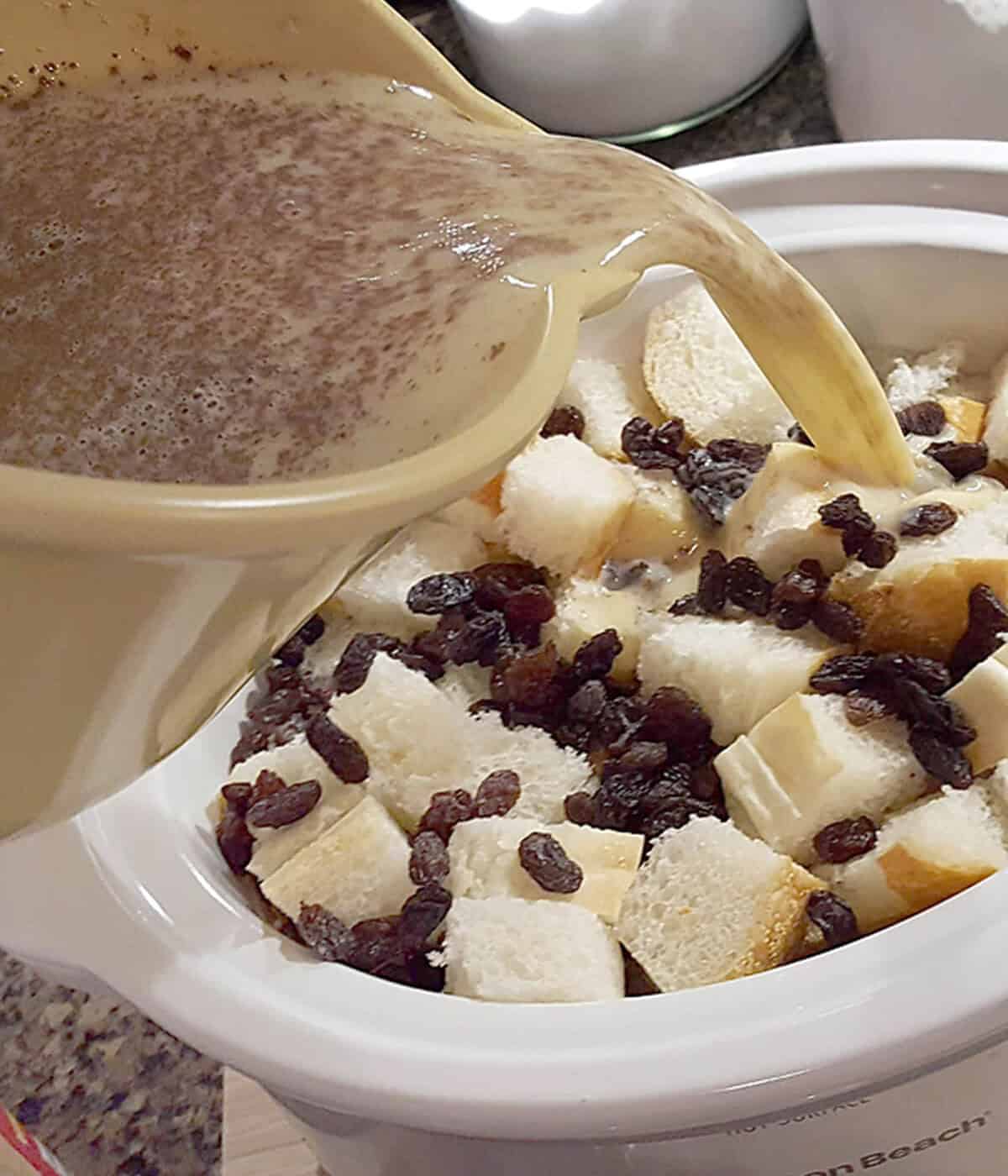 A bowl of liquid is being poured over bread cubes and raisins in a slow cooker.