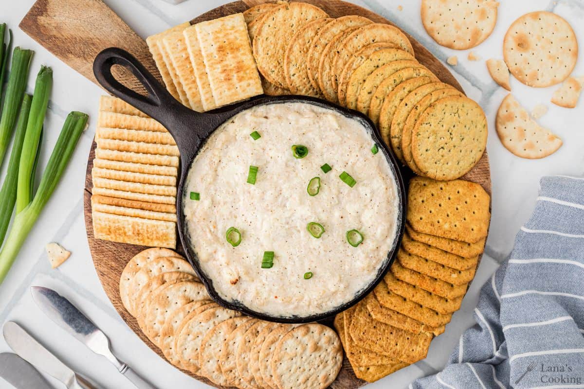 Round wooden board with creamy dip topped with green onions, surrounded by assorted crackers.