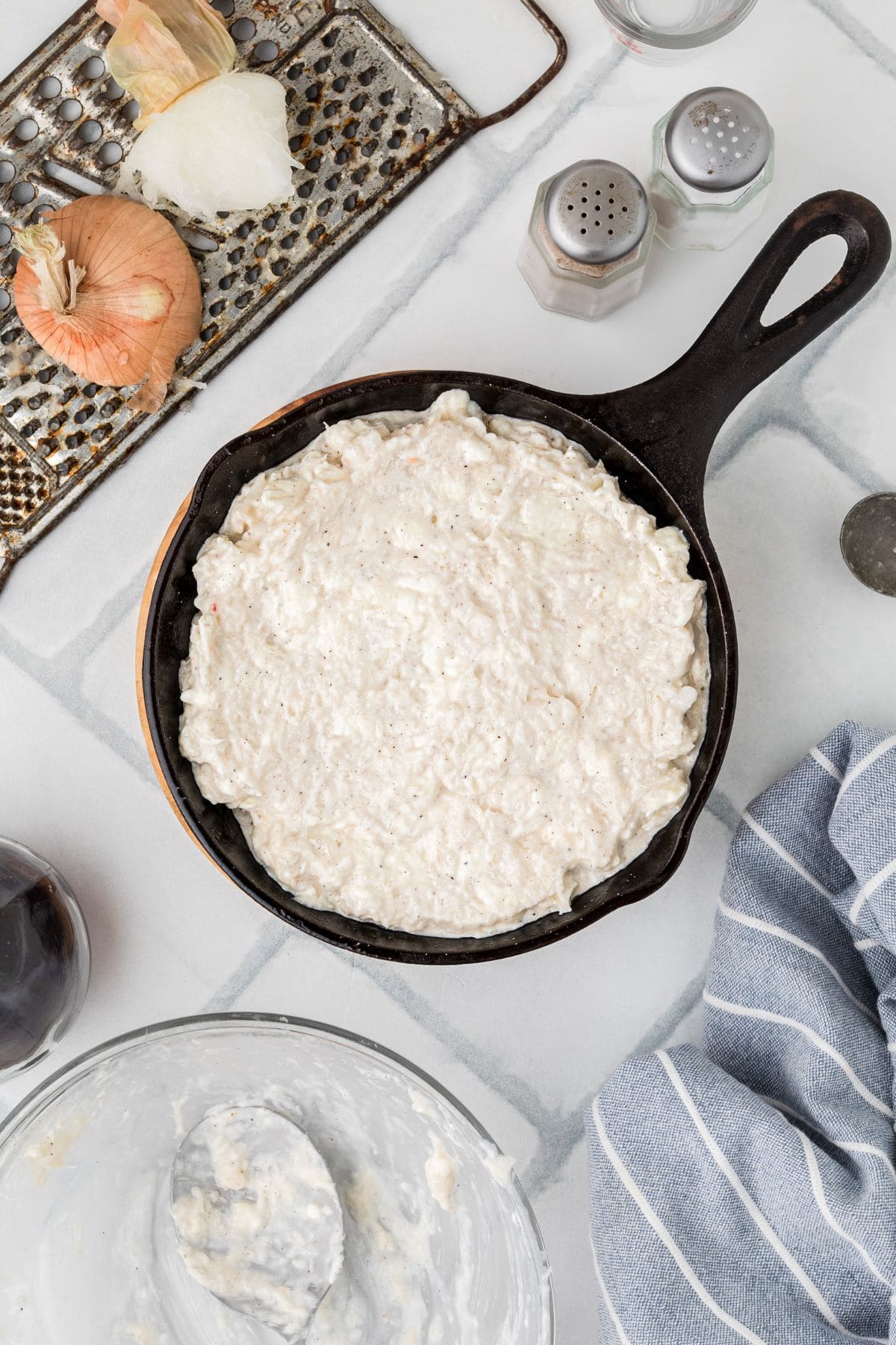 A cast iron skillet with a creamy white mixture, surrounded by onions, spices, and a blue-striped towel.