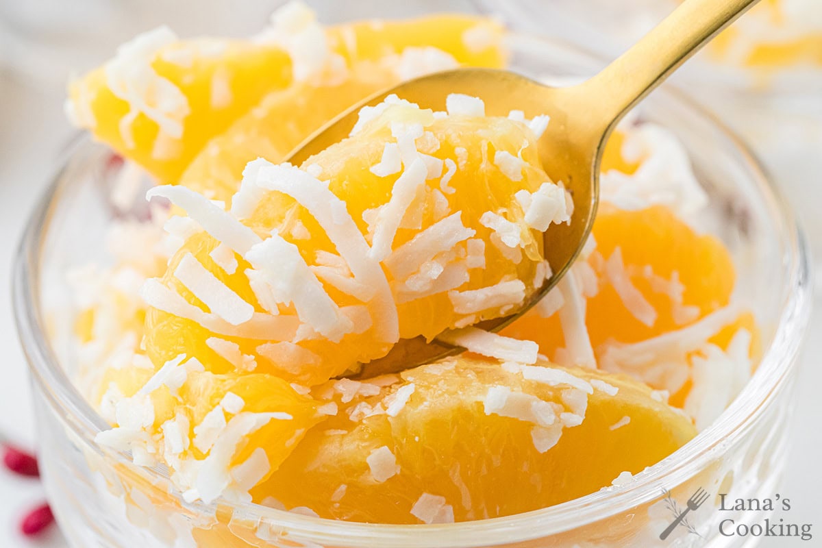 Close-up of orange slices topped with shredded coconut in a glass bowl, with a gold spoon.