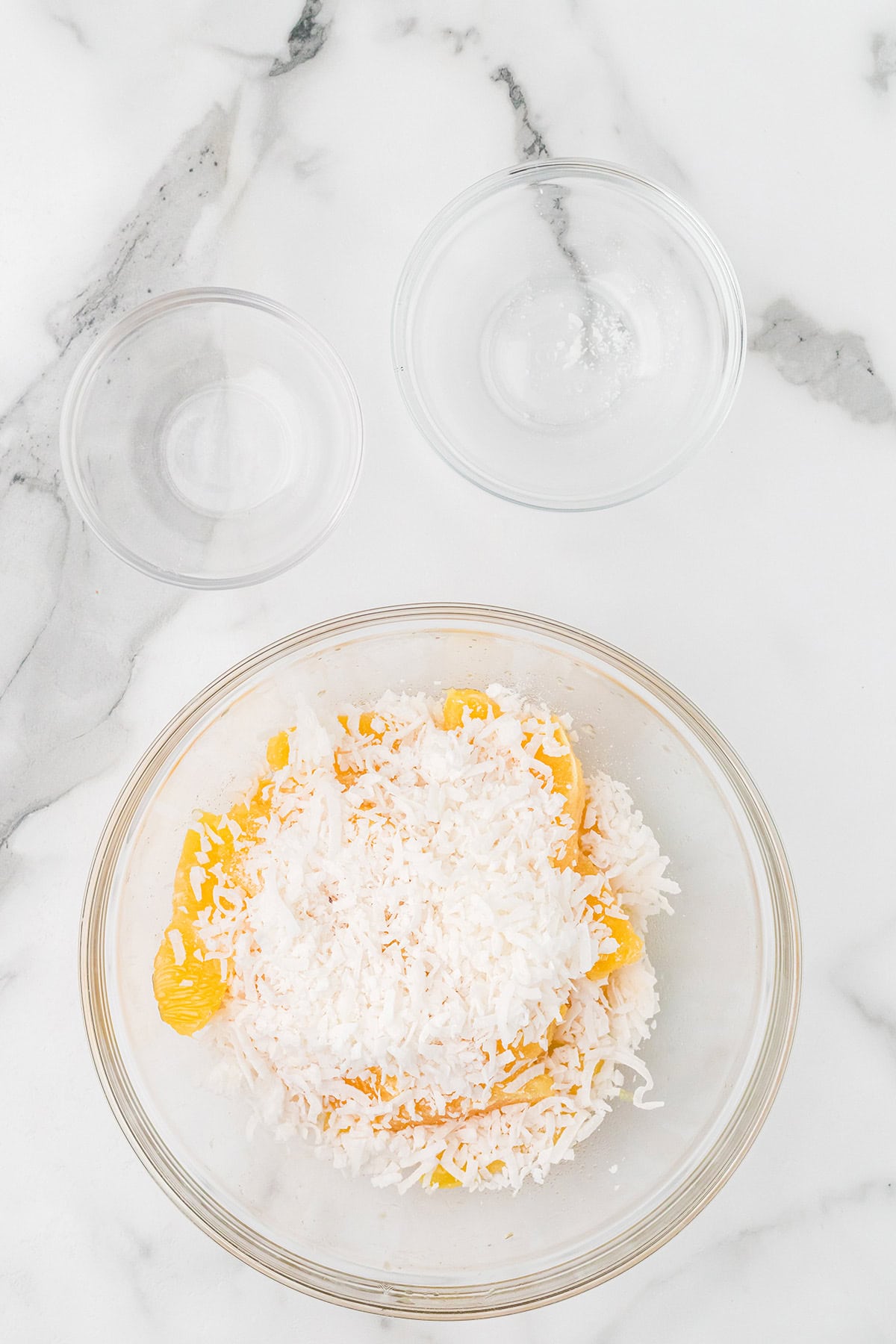 A bowl of orange segments and shredded coconut beside two glass bowls on a marble surface.
