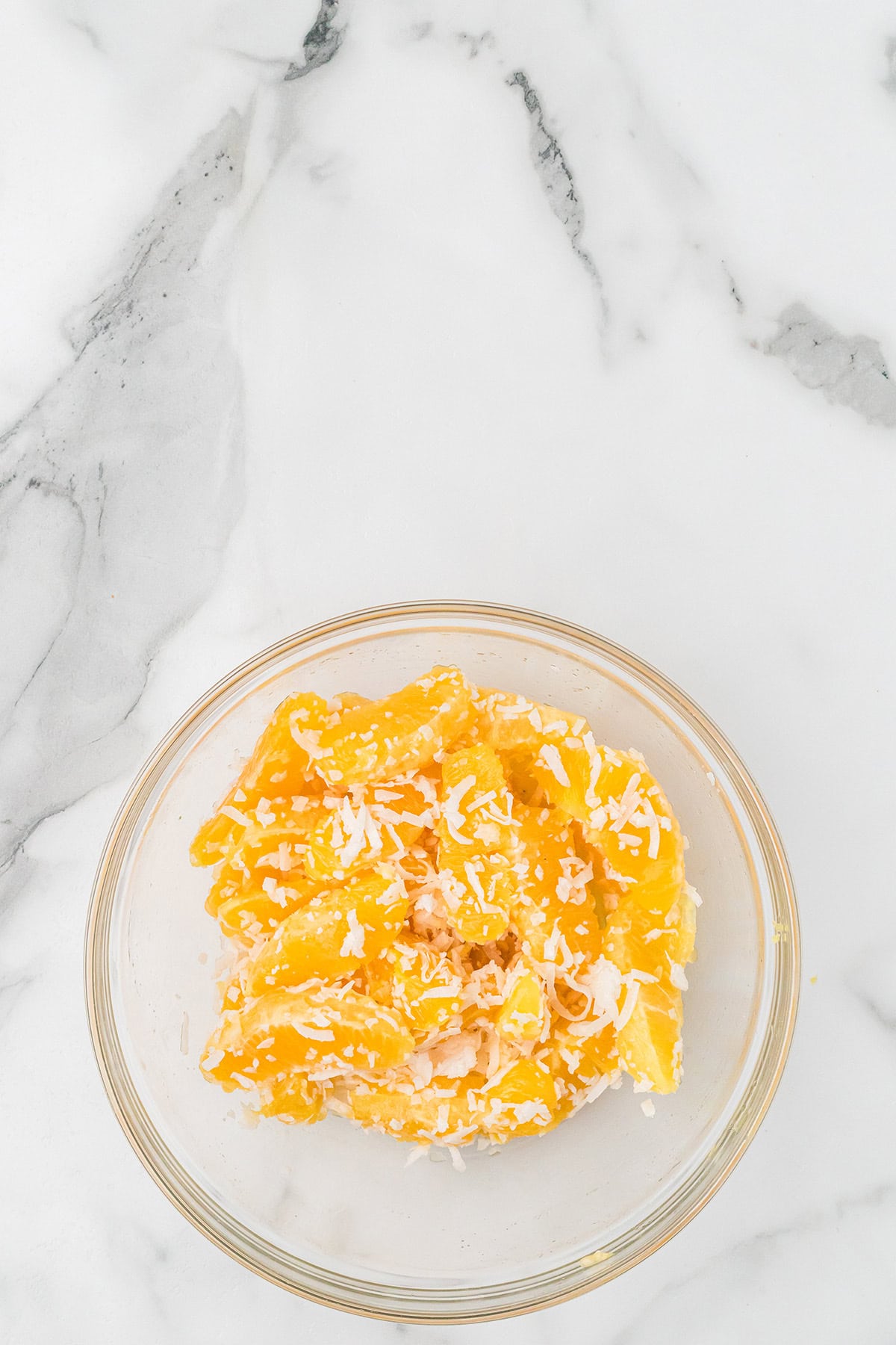 A glass bowl of orange slices topped with shredded coconut on a white marble surface.