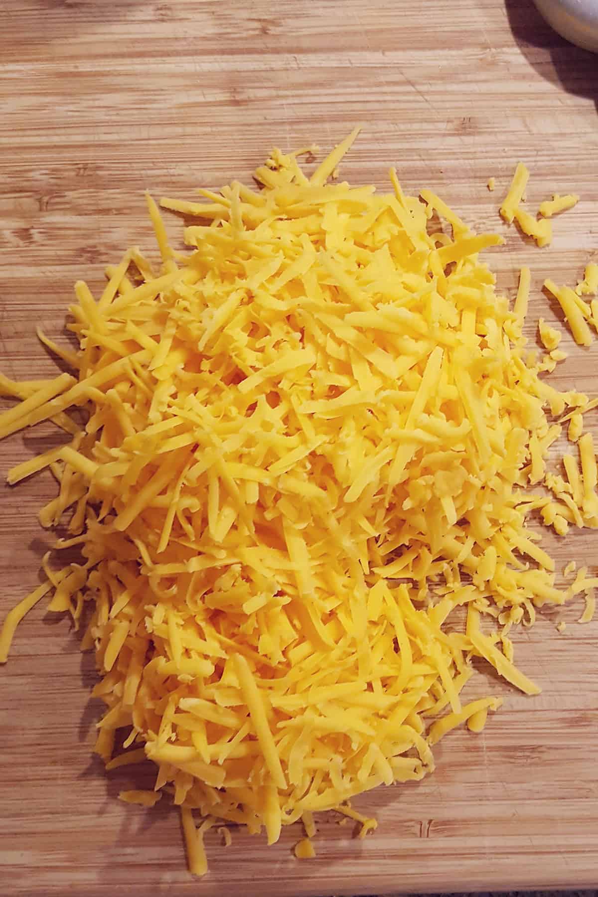 A pile of shredded yellow cheddar cheese on a wooden cutting board.