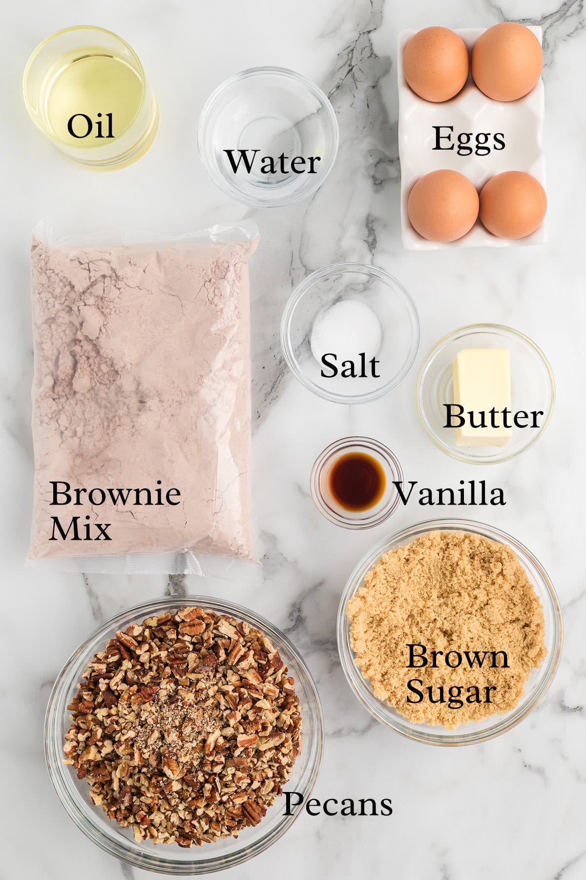 Brownie mix, oil, water, eggs, butter, vanilla, salt, brown sugar, and chopped pecans on a marble surface.