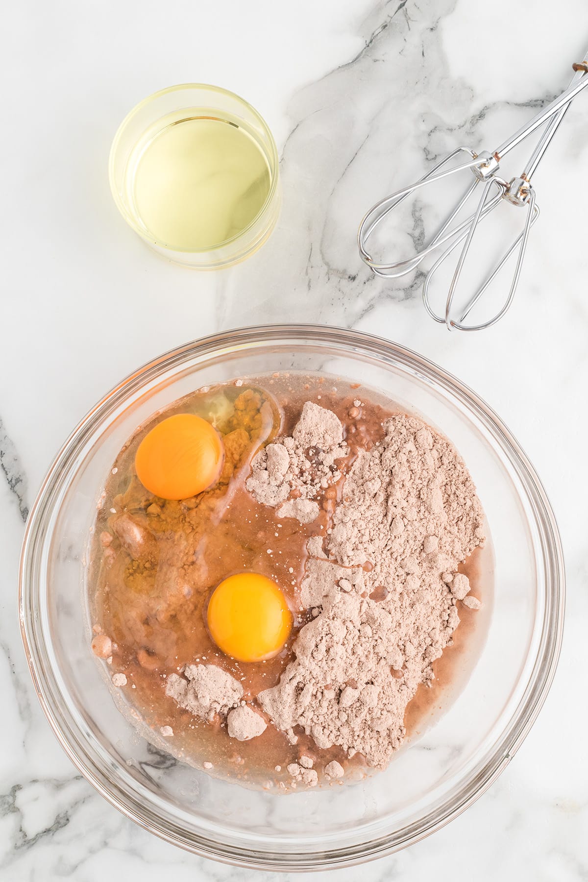 Glass bowl with brownie mix, eggs, and liquid, next to a cup of oil and an electric hand mixer on marble surface.