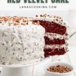 A three-layer red velvet cake with white frosting and chopped pecans on top, displayed on a cake stand with a slice being served.