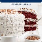 A sliced red velvet cake with cream cheese frosting and pecans on a white cake stand, with a plate of pecans nearby.