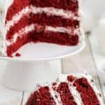 A slice of red velvet cake with white frosting sits on a plate in front of a cake stand holding the remaining cake.