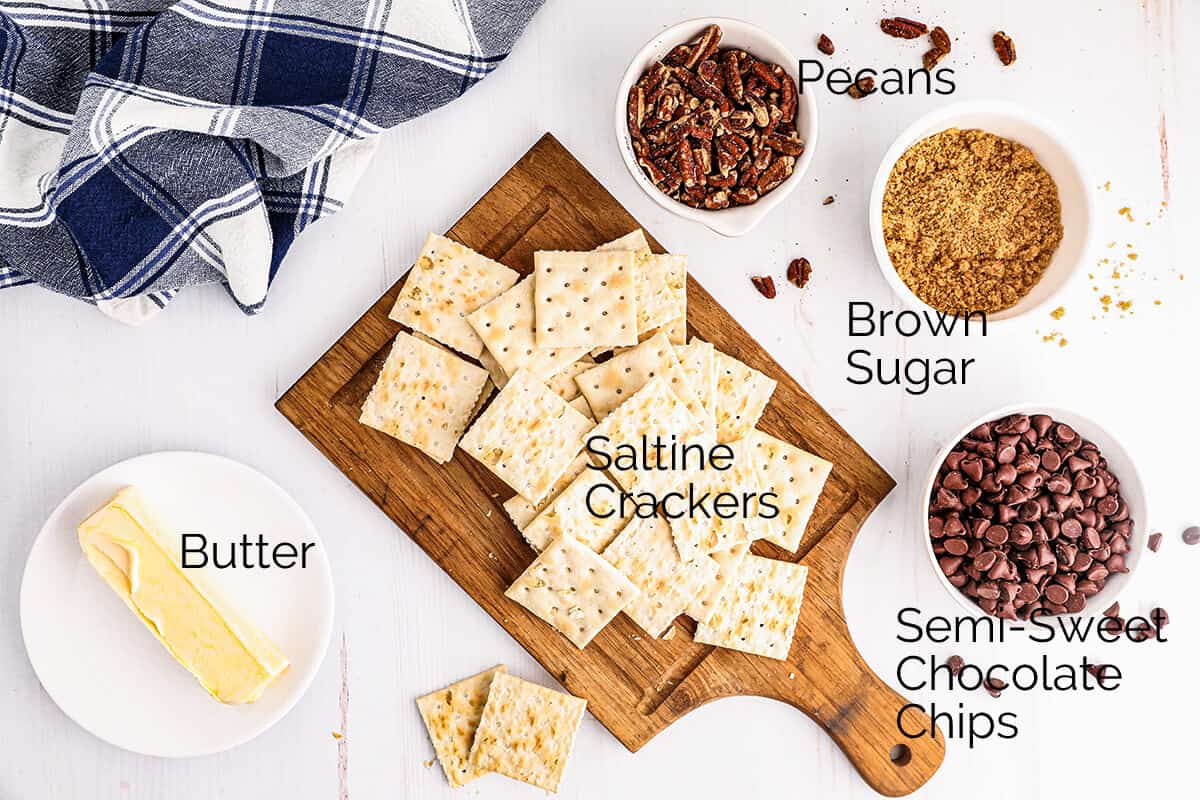 Flat lay of butter, saltine crackers, pecans, brown sugar, and chocolate chips labeled on a white table with a towel.