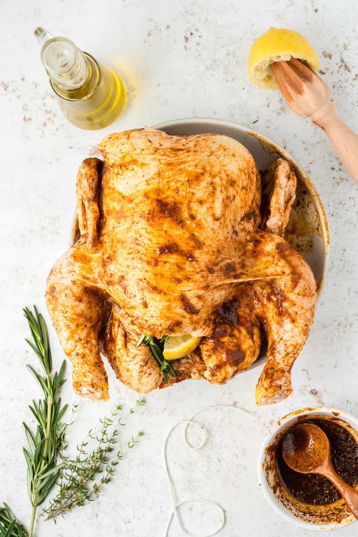 Whole seasoned raw chicken on a plate with herbs, lemon, oil, and marinade on a white surface.