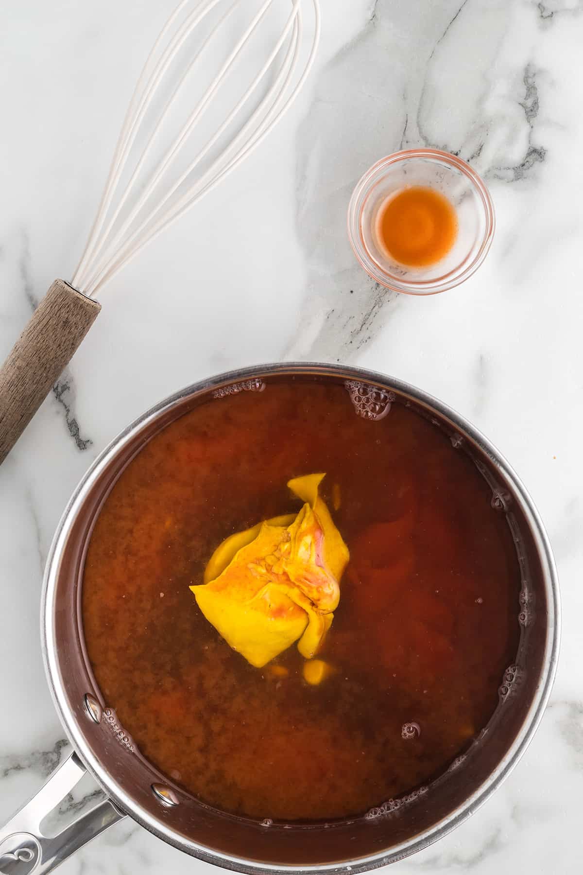 A saucepan filled with ingredients for barbecue sauce, next to a small bowl of liquid and a whisk on a marble surface.