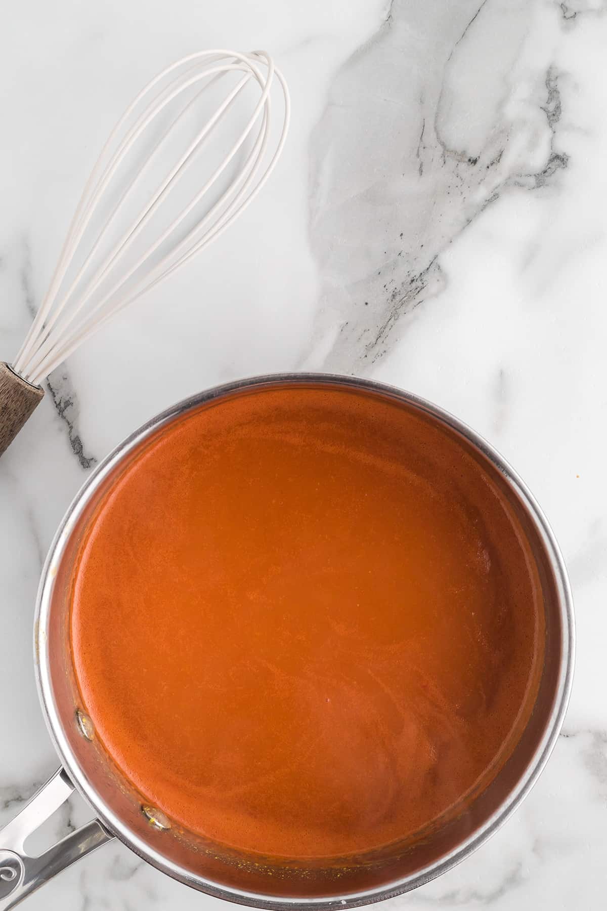 A saucepan filled with smooth barbecue sauce sits on a marble countertop beside a metal whisk.