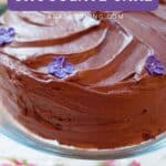 A chocolate cake with chocolate frosting and purple flower decorations on a glass stand.