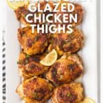 Eight glazed chicken thighs garnished with lemon slices and herbs on a white platter.