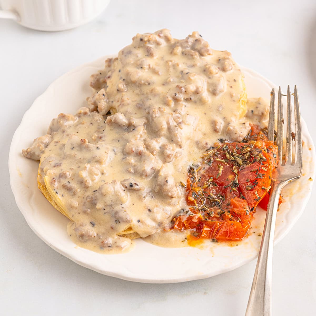 Biscuits topped with sausage gravy and a slice of seasoned roasted tomato on a white plate with a fork.