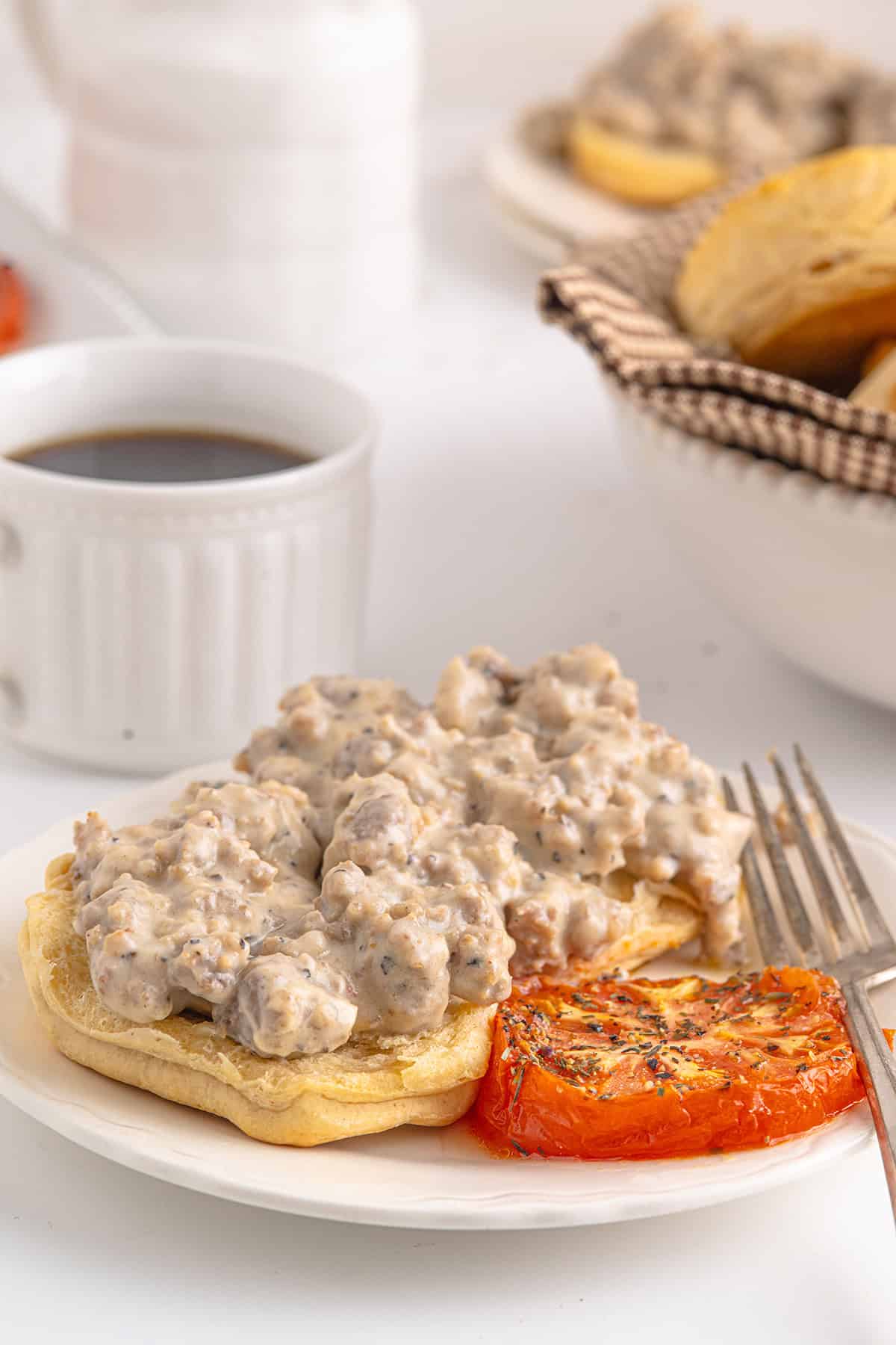 Open biscuit topped with sausage gravy, served with roasted tomato on a white plate; coffee cup in background.