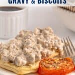 Open biscuits topped with sausage gravy, roasted tomato slices, and a fork on a plate, coffee cup in background.