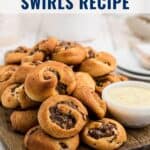 A pile of baked sausage swirls on a wooden board next to a bowl of dipping sauce.