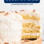 Four-layer coconut cake with sour cream frosting and pecan pieces, sitting on a wooden cake stand.