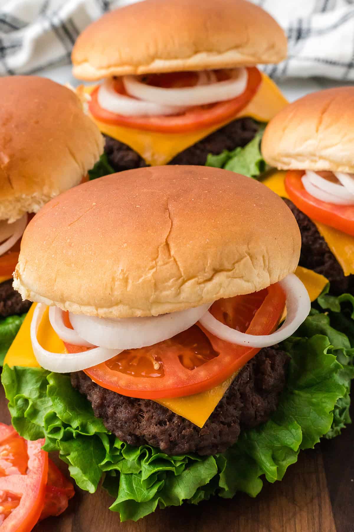 Four cheeseburgers with lettuce, tomato, onion, and cheddar cheese on buns, arranged on a wooden board.