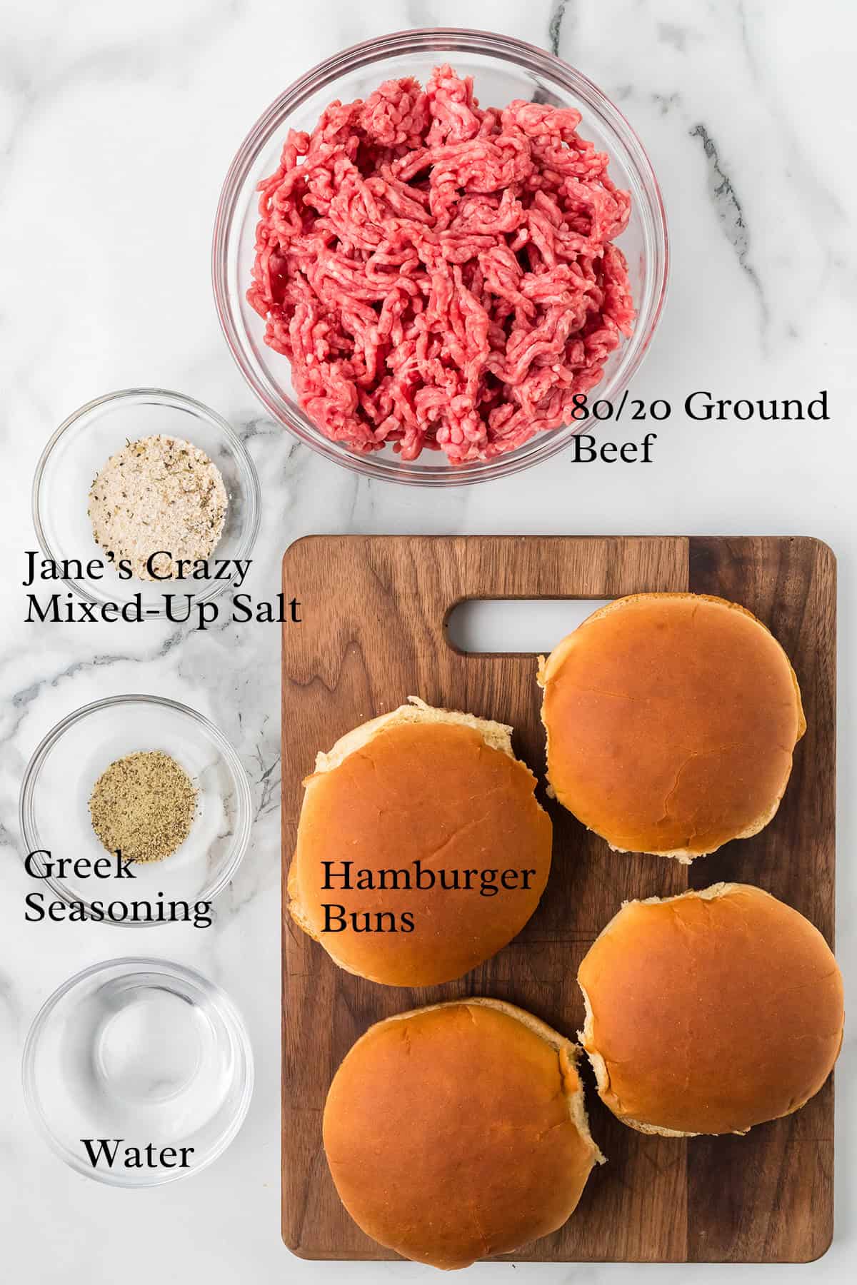Top view of ground beef, seasoning, water in bowls, and four hamburger buns on a marble and wood surface.