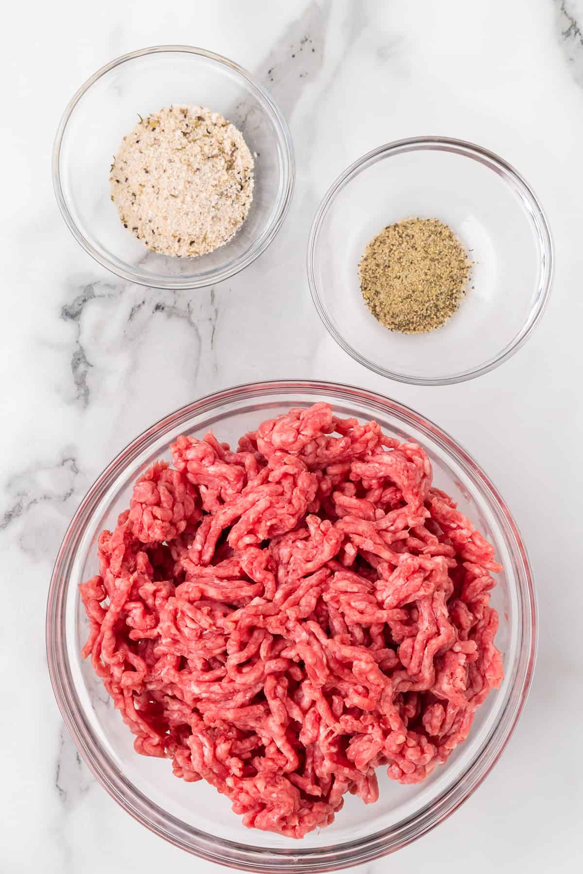 Three glass bowls with ground beef, breadcrumbs, and seasoning on a white marble surface.