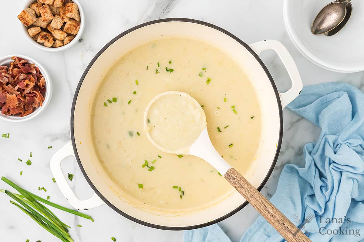 Creamy soup in a white pot with a ladle, garnished with chives, surrounded by bacon, croutons, and a blue napkin.
