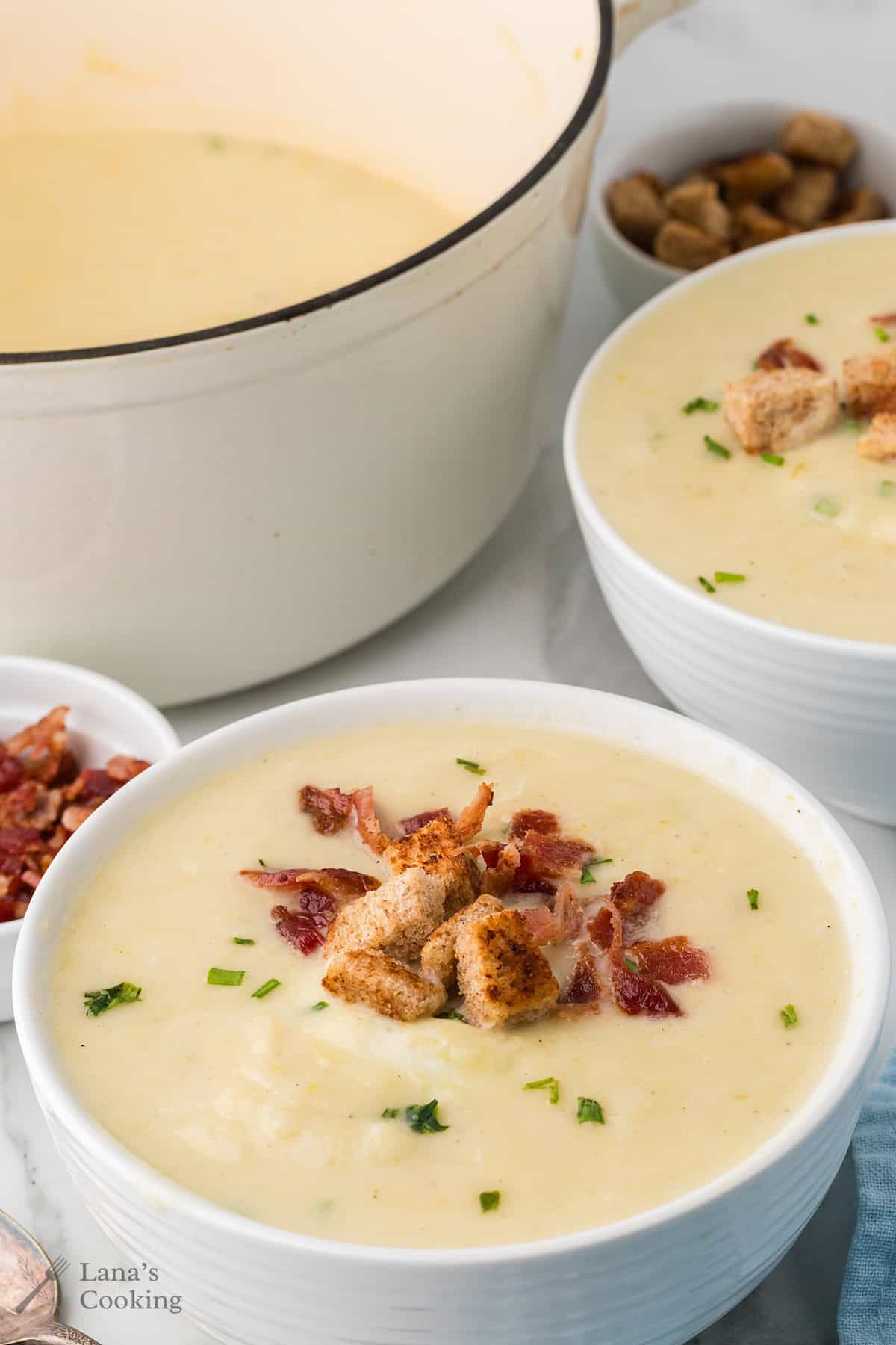 Two bowls of creamy soup topped with croutons and bacon, with a pot of soup and bacon pieces in the background.
